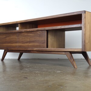 Puede incluir: Un centro de entretenimiento moderno de estilo mediados de siglo hecho de madera de nogal, con un caj&oacute;n y estantes abiertos. El centro tiene un estante de acento naranja brillante y patas inclinadas.