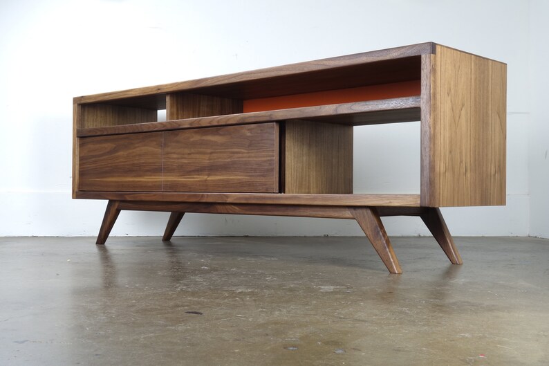 Puede incluir: Un centro de entretenimiento moderno de estilo mediados de siglo hecho de madera de nogal, con un caj&oacute;n y estantes abiertos. El centro tiene un estante de acento naranja brillante y patas inclinadas.