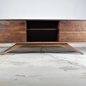 Puede incluir: Un centro de entretenimiento de madera de nogal de estilo moderno de mediados de siglo con dos puertas correderas y un estante. El gabinete tiene un acabado marr&oacute;n oscuro y se asienta sobre cuatro patas anguladas.