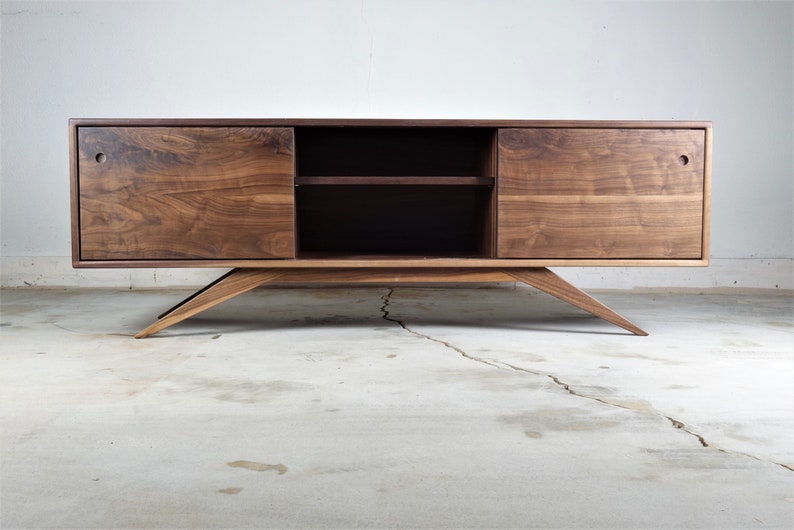 Puede incluir: Un centro de entretenimiento de madera de nogal de estilo moderno de mediados de siglo con dos puertas correderas y un estante. El gabinete tiene un acabado marr&oacute;n oscuro y se asienta sobre cuatro patas anguladas.