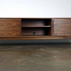 May include: A mid-century modern walnut wood entertainment center with two doors and a shelf. The doors have a wood grain pattern and the legs are angled.