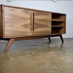 May include: A mid-century modern style walnut wood entertainment center with two doors and open shelving. The cabinet has angled legs and a light wood finish.