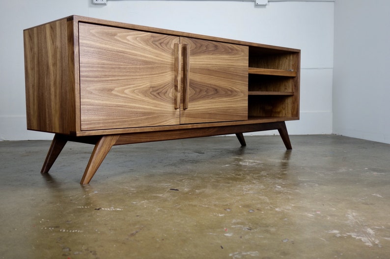 May include: A mid-century modern style walnut wood entertainment center with two doors and open shelving. The cabinet has angled legs and a light wood finish.
