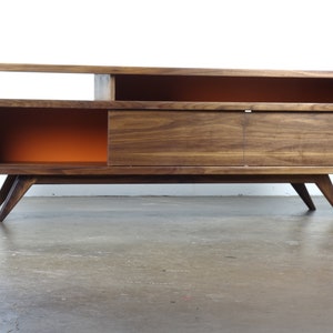 Puede incluir: Un centro de entretenimiento de madera de nogal de estilo moderno de mediados de siglo con una puerta corredera, estantes abiertos y paneles traseros pintados de naranja. La unidad tiene patas inclinadas y est&aacute; sobre un suelo de hormig&oacute;n.