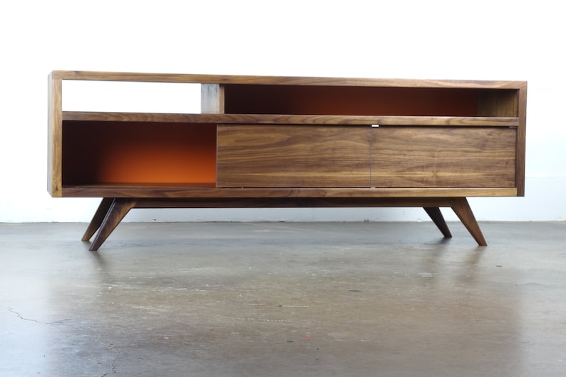 Puede incluir: Un centro de entretenimiento de madera de nogal de estilo moderno de mediados de siglo con una puerta corredera, estantes abiertos y paneles traseros pintados de naranja. La unidad tiene patas inclinadas y est&aacute; sobre un suelo de hormig&oacute;n.