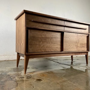 May include: A mid-century modern walnut wood sideboard with two sliding doors and a drawer. The sideboard has a rich brown color and a sleek design.