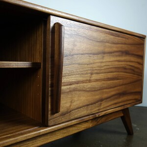 May include: A wooden entertainment center with a single door and a shelf. The door is made of dark wood with a grain pattern and has a simple, rectangular handle. The entertainment center has a light wood base and legs.