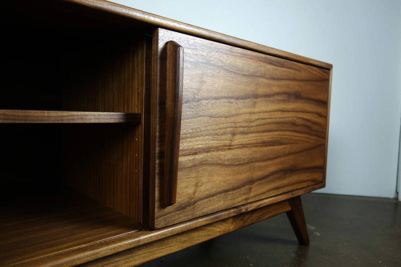 May include: A wooden entertainment center with a single door and a shelf. The door is made of dark wood with a grain pattern and has a simple, rectangular handle. The entertainment center has a light wood base and legs.