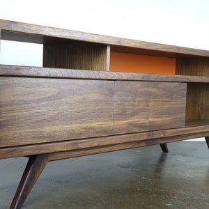 Puede incluir: Un centro de entretenimiento de estilo moderno de mediados de siglo en madera de nogal con una puerta corredera, estantes abiertos y un panel de acento naranja brillante. El centro de entretenimiento tiene cuatro patas.
