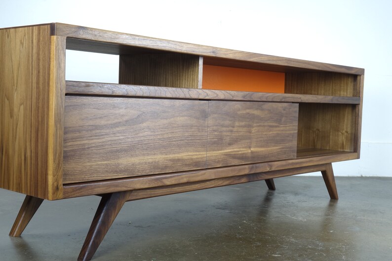 Puede incluir: Un centro de entretenimiento de estilo moderno de mediados de siglo en madera de nogal con una puerta corredera, estantes abiertos y un panel de acento naranja brillante. El centro de entretenimiento tiene cuatro patas.