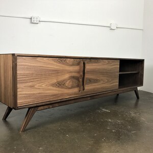 May include: A mid-century modern style walnut wood media console with two sliding doors and a shelf. The console has a dark brown finish and stands on four angled legs.