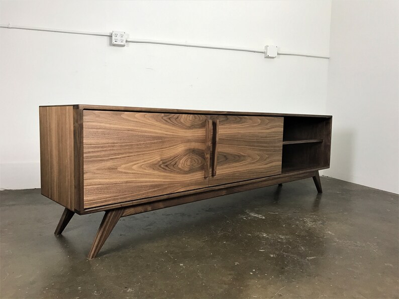May include: A mid-century modern style walnut wood media console with two sliding doors and a shelf. The console has a dark brown finish and stands on four angled legs.