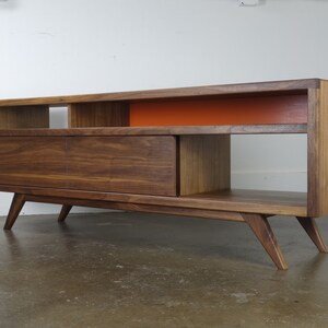 Puede incluir: Un centro de entretenimiento de estilo moderno de mediados de siglo en madera de nogal con un caj&oacute;n, estantes abiertos y un panel de acento naranja brillante. La unidad tiene patas inclinadas.