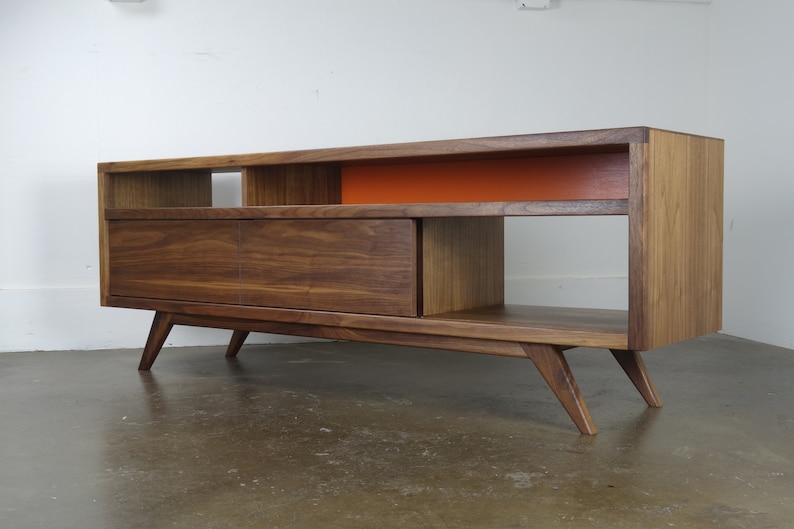 Puede incluir: Un centro de entretenimiento de estilo moderno de mediados de siglo en madera de nogal con un caj&oacute;n, estantes abiertos y un panel de acento naranja brillante. La unidad tiene patas inclinadas.