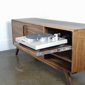 May include: A walnut wood record player stand with a sliding shelf for a turntable. The stand has two doors and a shelf inside. The turntable is a silver color and is sitting on the sliding shelf.