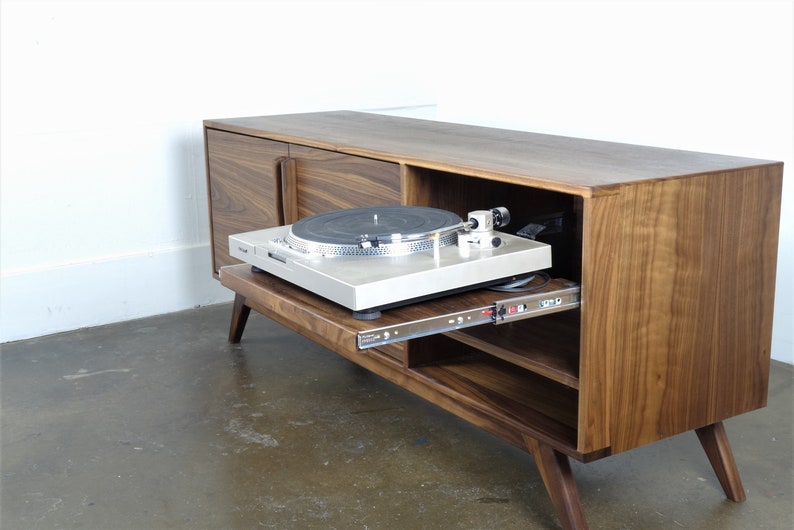 May include: A walnut wood record player stand with a sliding shelf for a turntable. The stand has two doors and a shelf inside. The turntable is a silver color and is sitting on the sliding shelf.