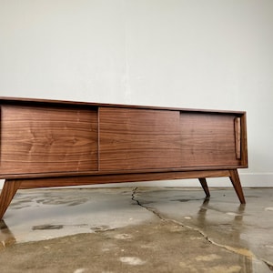 May include: A mid-century modern style wooden cabinet with sliding doors. The cabinet is a warm brown color and has angled legs. The doors have vertical handles. The cabinet is approximately 60 inches long.