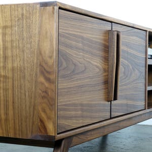 May include: A mid-century modern walnut wood entertainment center with two doors and open shelving. The doors have a unique, vertical, wood handle.