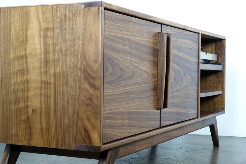 May include: A mid-century modern walnut wood entertainment center with two doors and open shelving. The doors have a unique, vertical, wood handle.