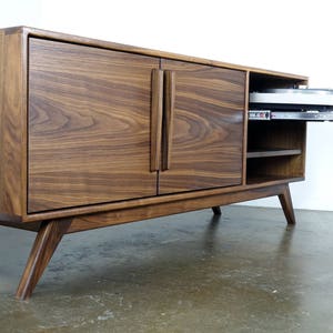 May include: A mid-century modern walnut wood entertainment center with two doors and a pull-out shelf. The doors have a vertical grain pattern and the legs are angled.