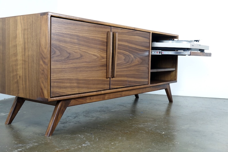 May include: A mid-century modern walnut wood entertainment center with two doors and a pull-out shelf. The doors have a vertical grain pattern and the legs are angled.