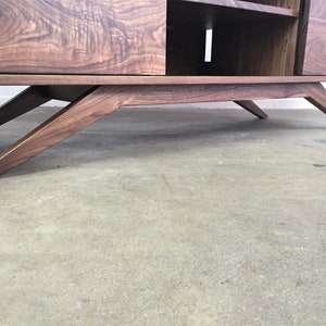 Puede incluir: Un centro de entretenimiento de estilo moderno de mediados de siglo en madera de nogal con un acabado en madera oscura y patas inclinadas. El centro de entretenimiento tiene dos puertas y estantes abiertos.