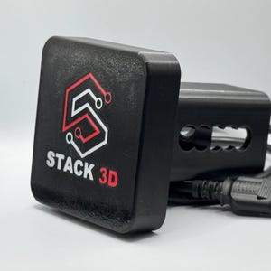 May include: Black rectangular device with the text "STACK 3D" in white and red. The device has a logo with a stylized "S" shape. A black cable and connector are attached to the device.
