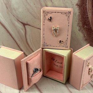 May include: Pink jewelry boxes shaped like books, with gold accents and heart and bow details. One box is open, revealing a silver ring with a dark blue stone. The inside of the box reads "All Love Wish You."