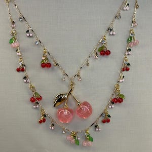 Op de afbeelding: Een gelaagde goudkleurige ketting met meerdere kleine kersenbedels met doorschijnend rood en roze fruit en groene blaadjes. Het middelpunt is een groter paar glinsterende roze kersen met een goudkleurig steeltje en een blad.