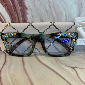 May include: Reading glasses with a dark tortoiseshell frame featuring decorative gold-tone starfish and blue-and-green turtle embellishments. The lenses are marked with a +2.25 magnification strength and blue light filtering technology.
