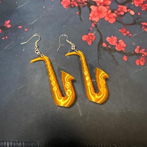 May include: A pair of gold-colored saxophone earrings featuring a detailed 3D-printed texture. Each earring is suspended from a silver-toned metal hook and displays the iconic curved shape of a saxophone with visible keys and a bell opening.