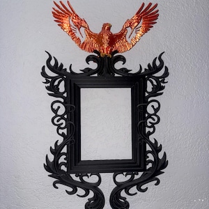 Puede incluir: Un marco de fotos rectangular con acabado negro mate y ornamentados remolinos en los laterales. Un fénix de color naranja metálico con las alas extendidas está montado en la parte superior central del marco, sobrepasando el borde.