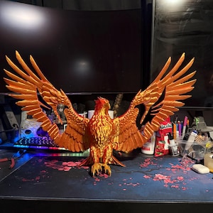 May include: A detailed, orange and gold phoenix sculpture with outstretched wings. The bird is perched on a desk, showcasing intricate feather details and a vibrant color scheme. The sculpture is a decorative item.