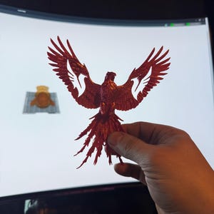 May include: A detailed, red-colored bird sculpture with outstretched wings. The bird has intricate feather detailing and a long, flowing tail. The sculpture is held in a hand, with a computer screen in the background.