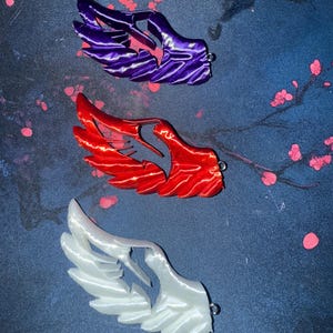 May include: Three wing-shaped pendants in purple, red, and white. Each wing has a detailed, layered design and a small loop for hanging. The pendants are displayed against a dark blue background with pink floral accents.
