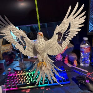 Può includere: Un ornamento a forma di uccello bianco stampato in 3D con ali spiegate e piume dettagliate. L'uccello ha un becco arancione e artigli color oro. È appeso a un filo verde, suggerendo che si tratti di un oggetto natalizio o decorativo.