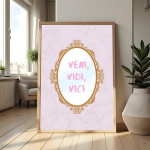 Puede incluir: Un cuadro enmarcado con la frase latina "VENI, VIDI, VICI" en rosa. El texto está dentro de un marco ovalado dorado, sobre un fondo rosa con vetas de mármol. El marco se exhibe sobre un suelo de madera.