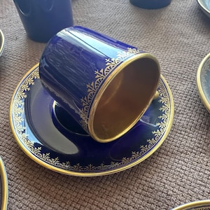 May include: A deep cobalt blue cup and saucer set featuring a metallic gold interior and a delicate gold filigree pattern along the rim of both pieces. The cup is positioned on its side on the saucer, showcasing the gold-toned interior finish.