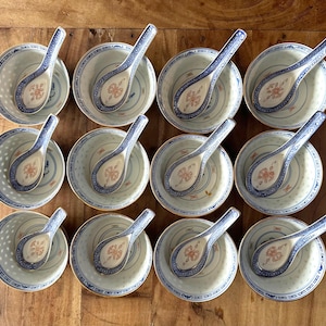 May include: A set of twelve white ceramic bowls and twelve matching ceramic spoons, each featuring a blue and white patterned rim and a central orange floral motif. The bowls have a translucent rice grain pattern around the sides.