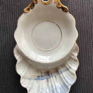 Puede incluir: Un plato de cerámica blanca con forma de concha marina y un asa con borde dorado en la parte superior. La sección inferior presenta un borde festoneado con detalles pintados en azul y gris, y el área superior tiene un interior blanco liso.
