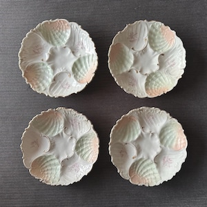 Puede incluir: Cuatro platos redondos de cerámica para ostras, cada uno con seis hendiduras en forma de concha. Los platos son blancos con suaves tonos pastel verde y rosa en las conchas, decorados con delicados motivos florales morados y bordes festoneados.