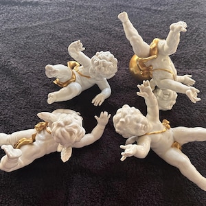 May include: Four white porcelain cherub figurines in various dynamic poses. Each cherub features detailed sculpted hair and is adorned with small, shiny gold-coloured accents around the waist or hips. The figures are arranged in a group, displaying different limb positions, including one figure positioned upside down and others reaching outward with arms and legs extended.