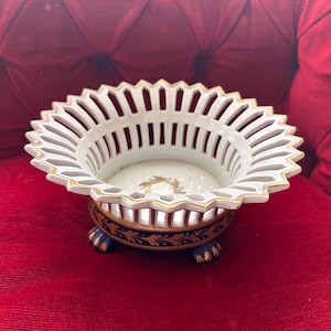 Puede incluir: Un cuenco de porcelana blanca con un borde festoneado tipo celosía y adornos dorados. La base es de color azul oscuro con patrones de hojas doradas y se apoya sobre tres pequeñas patas en forma de garra. Un diseño de corona dorada decora el centro.