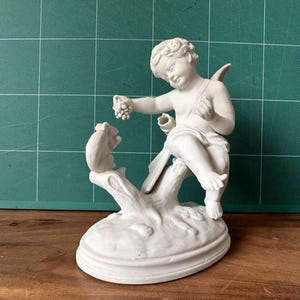May include: A matte white porcelain figurine depicting a cherub sitting on a tree stump, holding a cluster of grapes in one hand while reaching toward a small rabbit perched on the opposite side of the stump, all set on an oval base.