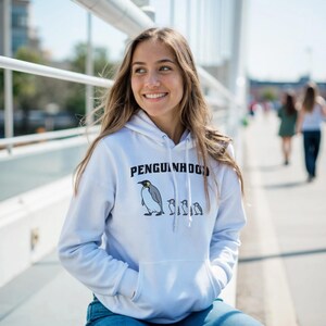 May include: White hoodie with the text "Penguinhood" above a cartoon penguin and three smaller penguins. The hoodie has a front pocket and a drawstring hood. The person is wearing blue jeans.