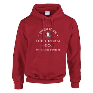 Op de afbeelding: Een rode hoodie met een witte grafische print op de borst. Het ontwerp bevat een kleine illustratie van een pinguïn, de tekst PENGUIN ICE CREAM CO. en de woorden WORLD'S FINEST, geaccentueerd met kleine hartjes en cirkelvormige symbolen.