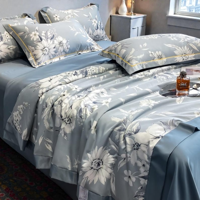 May include: A bed set featuring a light blue comforter and pillowcases with a large white floral pattern. The bedding has a solid light blue sheet and a gold trim on the pillowcases.