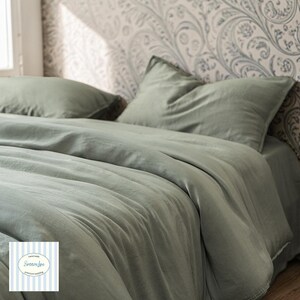 May include: A sage green bed set, comprising a duvet cover and two pillowcases, displayed on a bed. The fabric has a soft, slightly creased texture. The backdrop features patterned wallpaper in grey and white tones.