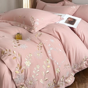 Może przedstawiać: Różowy haftowany zestaw pościeli z kwiatowymi wzorami. Zestaw zawiera poszwę na kołdrę i poszewki na poduszki. Haft przedstawia złote i srebrne wzory kwiatowe. Butelka perfum leży na kołdrze.