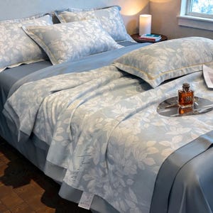 May include: A light blue bed set with a white floral pattern. The set includes pillows, a comforter, and a fitted sheet. A silver tray with a perfume bottle and a small knife rests on the comforter. The bed is in a well-lit room.